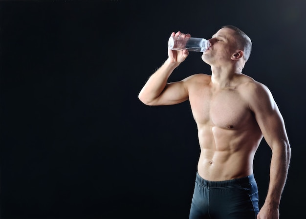 Jonge gespierde man drinkt water uit een doorzichtige plastic fles op een donkere studioachtergrond met achtergrondverlichting.