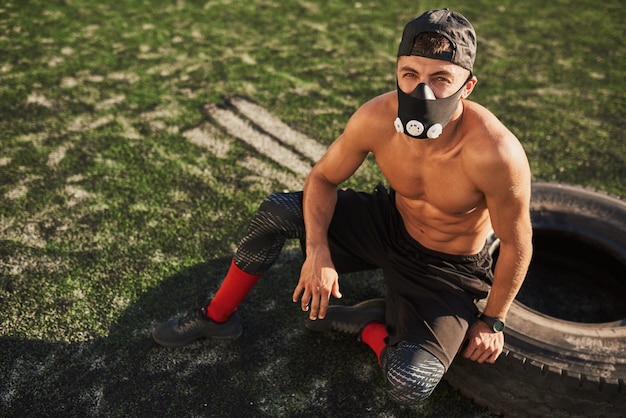 Jonge gespierde man zit in masker om de belasting van de ademhalingsspieren te vergroten na het zitten op de band na het sporten buitenshuis Ruimte voor uw tekst kopiëren Shirtless sportman ontspannen na het sporten in het stadion