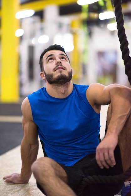 jonge gespierde man zitten en ontspannen voor touw klimmen in cross fitness gym