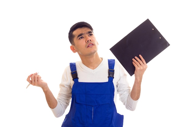 Jonge gestoorde man met donker haar in een wit overhemd en een blauwe overall met witte pen en zwarte map en denkend op een witte achtergrond in de studio