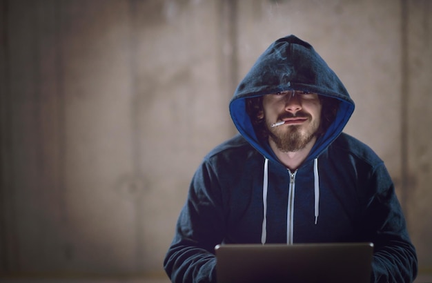 Jonge getalenteerde hacker die een sigaret rookt terwijl hij werkt op een laptopcomputer in een donker kantoor met een betonnen muur op de achtergrond