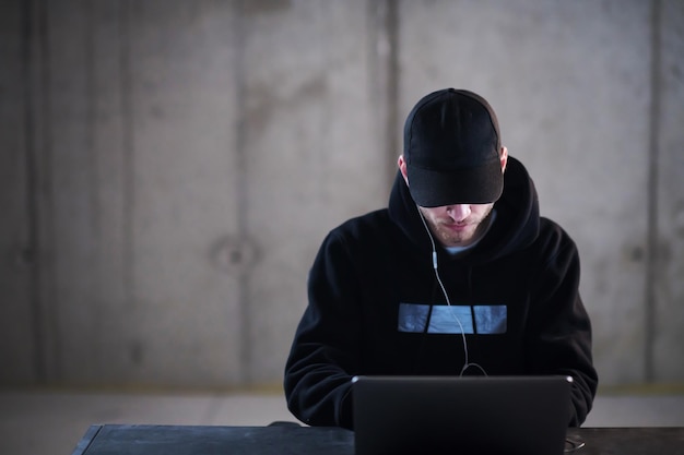 Jonge getalenteerde hacker die laptopcomputer gebruikt terwijl hij in een donker kantoor werkt met een betonnen muur op de achtergrond