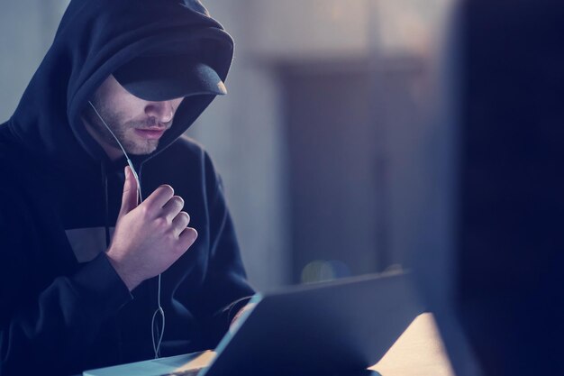 Jonge getalenteerde hacker die laptopcomputer gebruikt terwijl hij in een donker kantoor werkt met een betonnen muur op de achtergrond