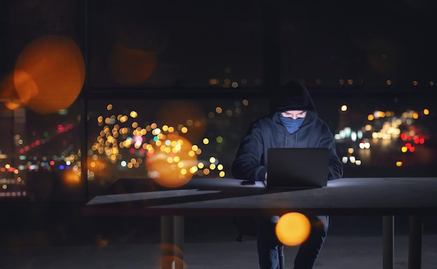 Jonge getalenteerde hacker die laptopcomputer gebruikt terwijl hij 's nachts in een donker kantoor werkt met grote stadslichten op de achtergrond