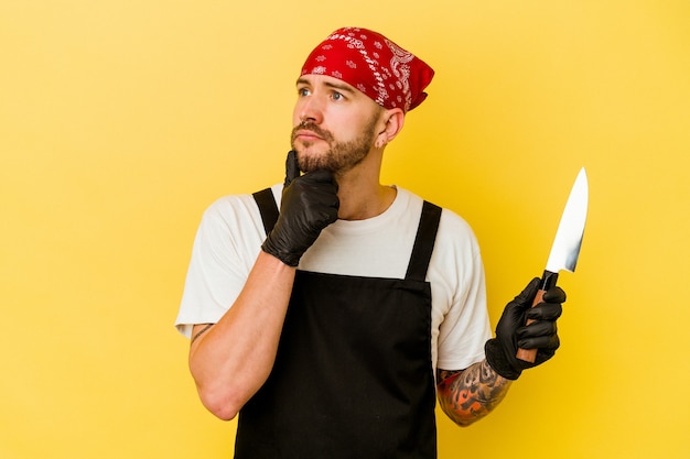 Jonge getatoeëerde batcher blanke man met een mes geïsoleerd op gele achtergrond opzij kijken met twijfelachtige en sceptische uitdrukking.