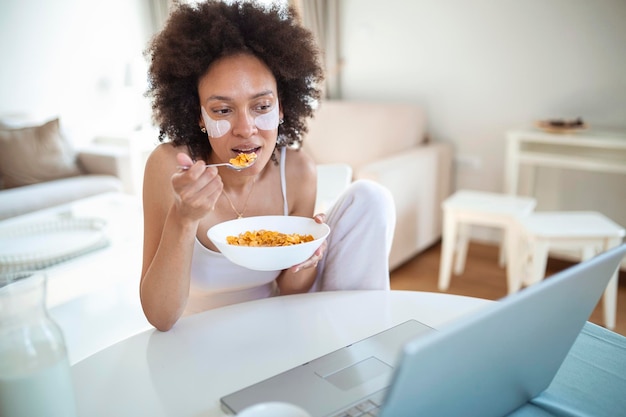 Jonge geweldige vrouw die binnen aan tafel zit met een laptop die cornflakes vasthoudt Kijkend naar een laptopcomputer en praat met haar vrienden via een videogesprek