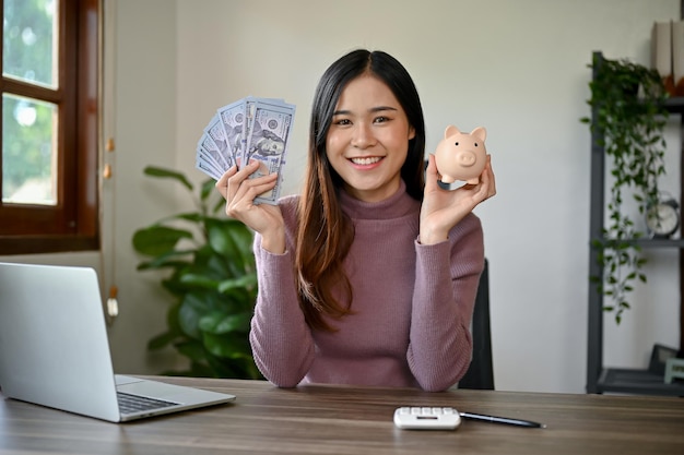 Jonge glimlachende Aziatische vrouw die haar geld bespaart met behulp van Piggy Bank