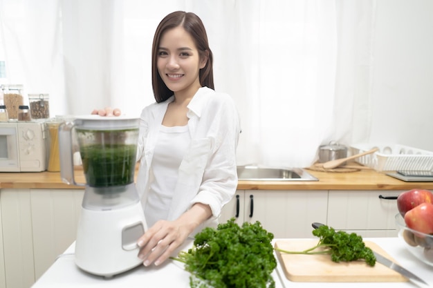 Jonge glimlachende Aziatische vrouw die smoothie maakt met verse groenten in de blender in minimalistische keuken