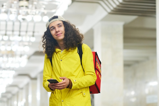 Jonge glimlachende bezorger in gele jas die bij het metrostation staat en vooruit kijkt