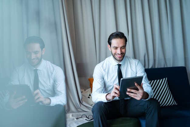 Jonge glimlachende blanke zakenman in formele slijtage met behulp van tablet voor werk en zittend op de bank in kantoor.