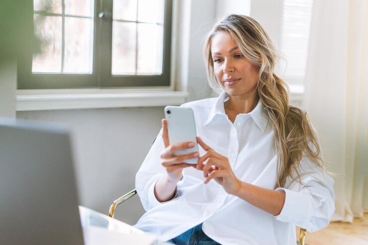 Jonge glimlachende blonde vrouw met lang haar in wit overhemd met smartphone in de heldere moderne kantoorvrouw stuurt een bericht per smartphone