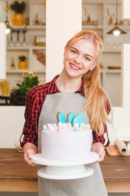 Jonge glimlachende blonde vrouwenchef-kok die een eigengemaakte verfraaide witte Pasen-cake met eieren houdt.