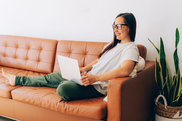 Jonge glimlachende brunette vrouw die aan haar laptop werkt en thuis op de bank zit.