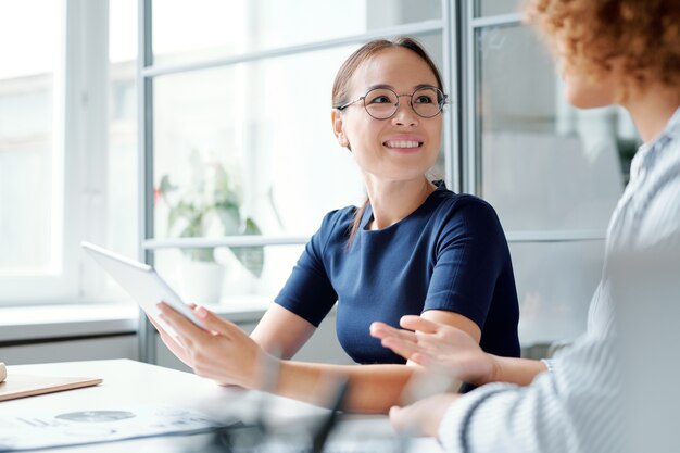 Jonge glimlachende elegante analist met touchpad die collega bekijkt terwijl hij naar haar mening over het onderwerp luistert