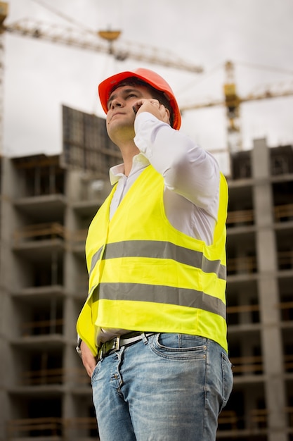 Jonge glimlachende ingenieur die telefonisch praat bij het gebouw in aanbouw