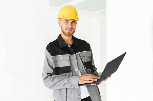 Foto jonge glimlachende ingenieur in werkkleding en gele veiligheidshelm die vreugdevol in de camera kijkt terwijl hij de laptop in de hand houdt op een witte achtergrond