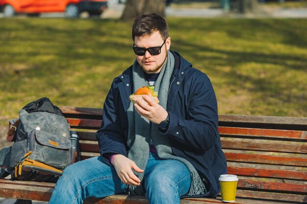 Jonge glimlachende man die hamburger eet en koffie drinkt terwijl hij op de stadsbank zit