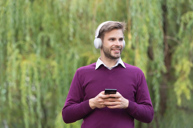 Jonge Glimlachende man luistert naar muziek in een koptelefoon en chat buiten op de telefoon