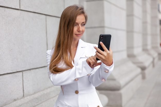 Jonge glimlachende onderneemster in witte laag die smartphone gebruiken