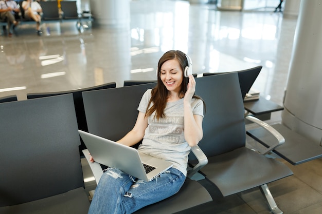 Jonge glimlachende reizigerstoeristenvrouw met koptelefoon die muziek luistert die op laptop werkt, wacht in de lobby op de internationale luchthaven