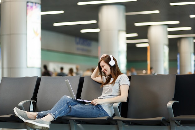 Jonge glimlachende reizigerstoeristenvrouw met koptelefoon die muziek luistert die op laptop werkt, wacht in de lobby op de internationale luchthaven