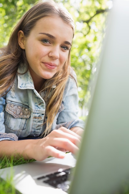 Jonge glimlachende student die op het gras ligt dat haar laptop met behulp van