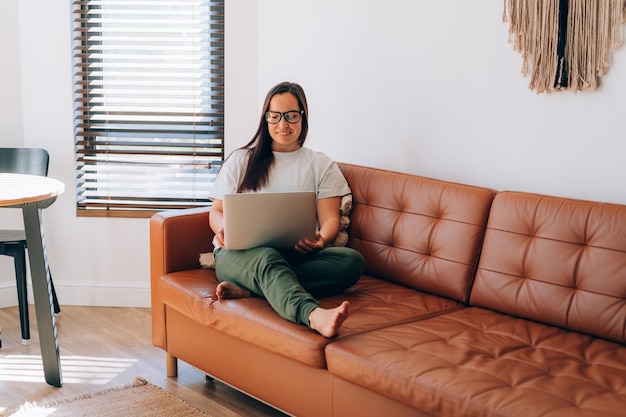 Jonge glimlachende vrouw die aan haar laptop werkt en thuis op de bank zit