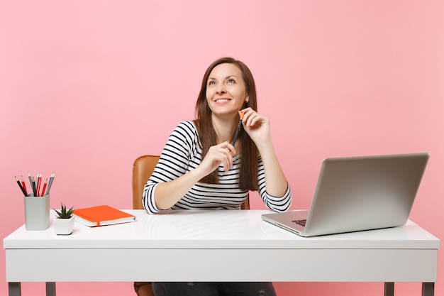 Jonge glimlachende vrouw die een potlood vasthoudt en opkijkt terwijl ze dromend zit te werken aan een wit bureau met een moderne pc-laptop