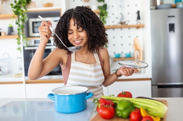 Jonge glimlachende vrouw die geniet van smaak en geur tijdens het koken van de lunch in de keuken