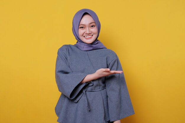 Jonge glimlachende vrouw die hijab draagt, toont iets en houdt iets vast met handpalmen geïsoleerd op een lichtgele bannerachtergrond