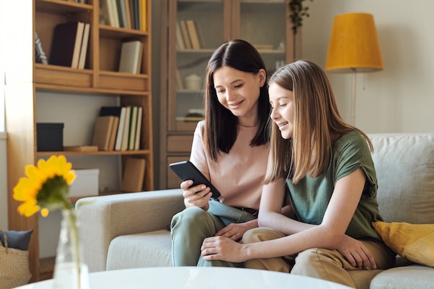 Jonge glimlachende vrouw die in smartphone door foto's scrolt terwijl ze ze aan haar schattige blonde tienerdochter in de woonkamer laat zien