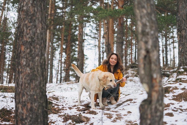 Jonge glimlachende vrouw in geel jasje met grote vriendelijke witte hond Labrador die in de winterbos loopt