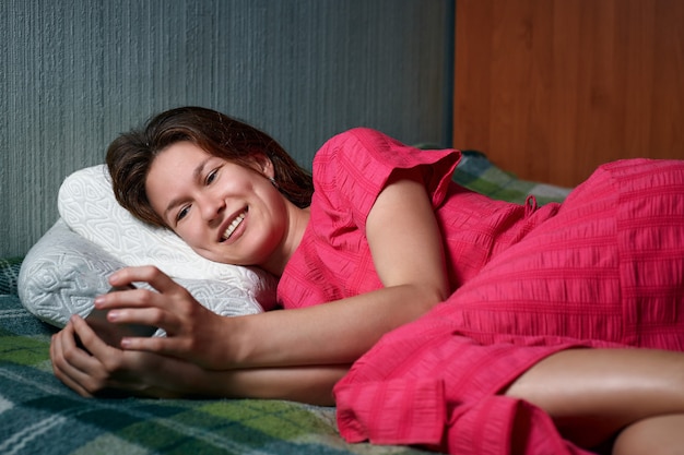 Jonge glimlachende vrouw in rode kleding die op het bed ligt en smartphone gebruikt