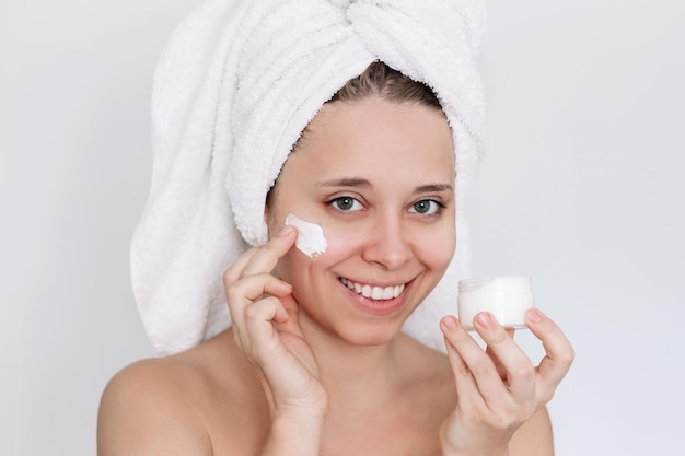 Jonge glimlachende vrouw met een handdoek op haar hoofd na de douche die een smeer van crème op de huid aanbrengt