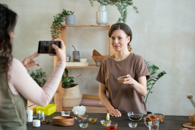 Foto jonge glimlachende vrouw met een kleine kom met essentieolie die naar de smartphonecamera kijkt die door haar vriend wordt vastgehouden terwijl ze bij de tafel staat