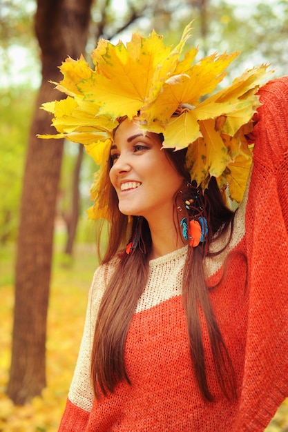 Jonge glimlachende vrouw met krans van esdoornbladeren op een hoofd, buitenportret in herfstpark