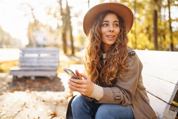 Jonge glimlachende vrouw zit op een bankje in het herfstpark met een telefoon in stijlvolle kleren in een gelukkige stemming