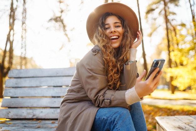 Jonge glimlachende vrouw zit op een bankje in het herfstpark met een telefoon in stijlvolle kleren in een gelukkige stemming