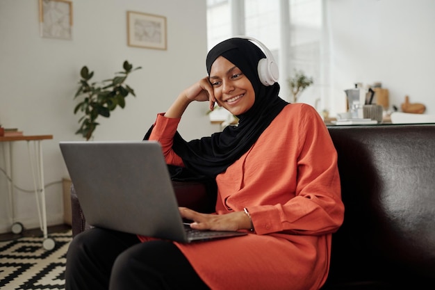 Jonge glimlachende zakenvrouw in hijab en casualwear die naar het scherm van een laptop kijkt