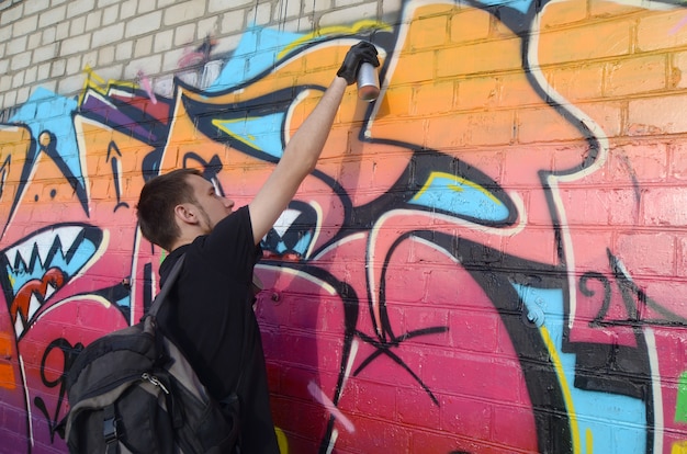 Jonge graffitikunstenaar met rugzak en gasmasker op zijn nek schildert kleurrijke graffiti