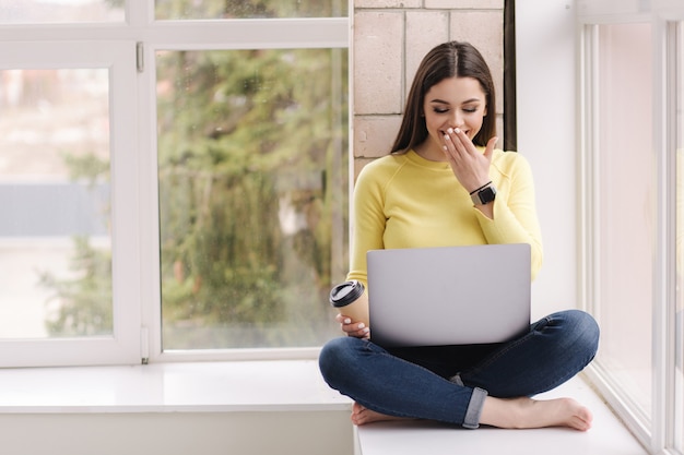 Jonge grafisch ontwerper thuis werken. Vrouw zittend op de vensterbank met laptop en koffie drinken.