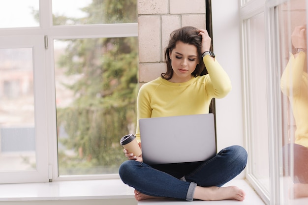 Jonge grafisch ontwerper thuis werken. vrouw zittend op de vensterbank met laptop en koffie drinken.