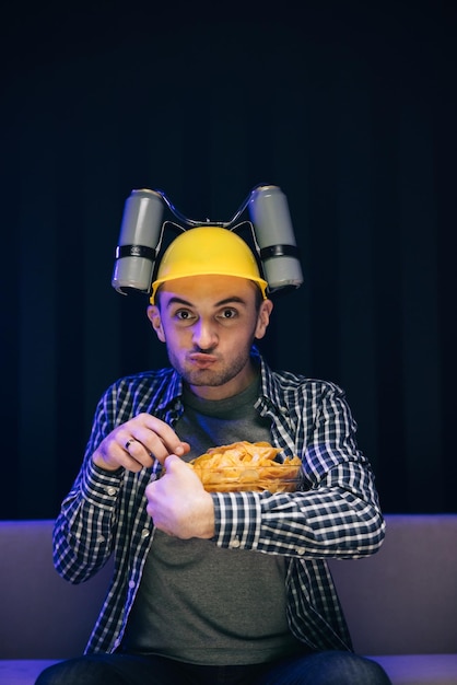 Jonge grappige man met bierhelm op het hoofd die chips eet terwijl hij naar een film kijkt op de bank in de woonkamer