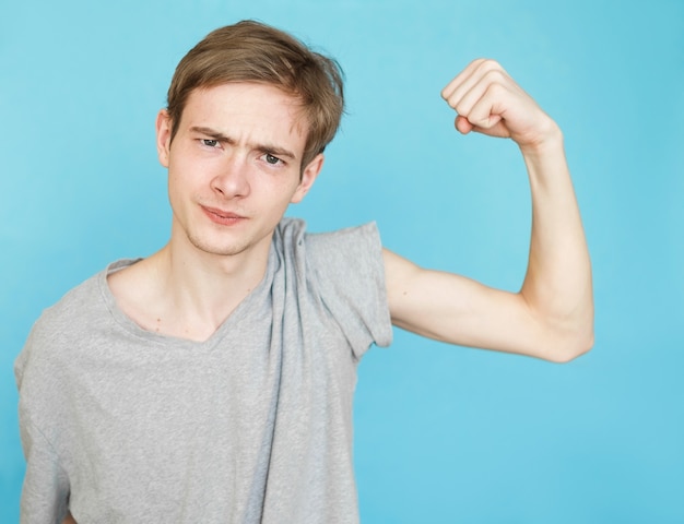 Foto jonge grappige mannelijke tiener in grijs t-shirt op blauwe achtergrond toont zijn spieren, na gewichtsverlies