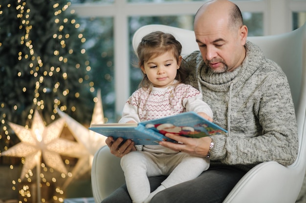 Jonge grootvader speelt en leest een boek met haar schattige kleindochter in de kerststemming van de dennenboom