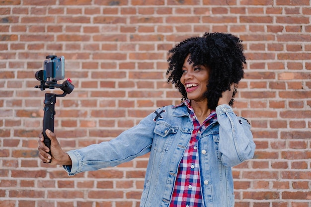 Jonge Haïtiaanse vrouw lachend met afrohaar bakstenen achtergrond die een vlog opneemt met een mobiele telefoon en gymbal