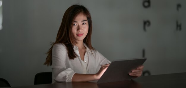 Jonge hardwerkende Aziatische onderneemster die aan haar project werkt terwijl het gebruiken van tablet in moderne bureauruimte bij nacht