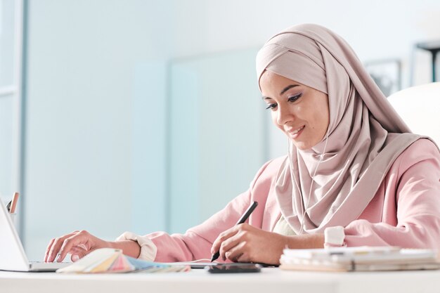 Jonge hedendaagse islamitische vrouwelijke ontwerper in hijab en oortelefoons die afbeeldingen retoucheren terwijl ze de stylus op het tabletscherm op kantoor houden