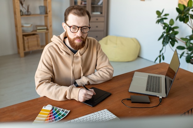 Jonge hedendaagse serieuze mannelijke freelance ontwerper in brillen en vrijetijdskleding met behulp van touchpad en stylus tijdens het retoucheren van afbeeldingen