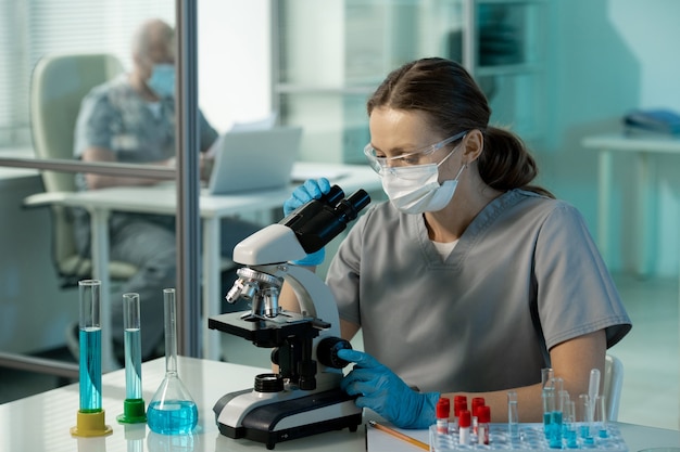 Jonge hedendaagse vrouwelijke wetenschapper in uniform, beschermend masker en bril die naar chemische monsters in de microscoop in het laboratorium kijkt